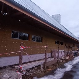 GAMB-POL - Budynek w trakcie ocieplania elewacji, widoczna warstwa izolacyjna z wełny mineralnej, częściowo osłonięta folią, okna zamontowane, teren budowy zabezpieczony taśmą ostrzegawczą, w tle las iglasty.