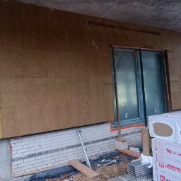 GAMB-POL - Ocieplanie ściany budynku wełną mineralną Rockwool, widoczne pakiety materiału na pierwszym planie, rusztowanie z tyłu, okno zabezpieczone folią, etap prac w toku.