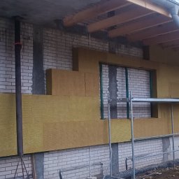 GAMB-POL - Budynek w trakcie docieplania: żółte płyty izolacyjne naklejone na fragment ściany z białej cegły, widoczne rusztowania i paczki materiałów izolacyjnych, okno częściowo zakryte izolacją.