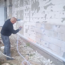 GAMB-POL - Pracownik w czarnej bluzie natryskuje białą substancję na szarą ścianę budynku z pustaków, używając pistoletu natryskowego z długim wężem; widoczne niedokończone fragmenty i surowy teren wokół.