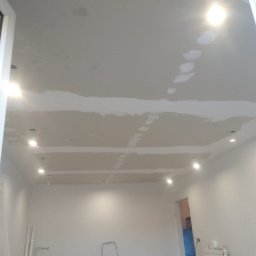 GAMB-POL - Sufit w trakcie prac wykończeniowych, widoczne płyty kartonowo-gipsowe, szpachlowane łączenia oraz otwory na oświetlenie punktowe, w tle drabina i materiały budowlane, w oddali widoczna osoba.