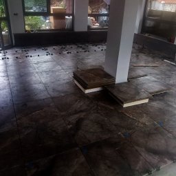 GAMB-POL - Układanie ciemnych, marmurkowych płytek podłogowych w jasnym pomieszczeniu z dużymi oknami. Widoczne narzędzia i akcesoria glazurnicze, płytki ułożone częściowo.