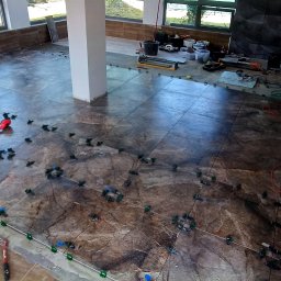 GAMB-POL - Układanie płytek podłogowych w Świebodzicach. Widoczne narzędzia, poziomice i system poziomowania płytek na brązowych płytkach z marmurowym wzorem w przestronnym wnętrzu.