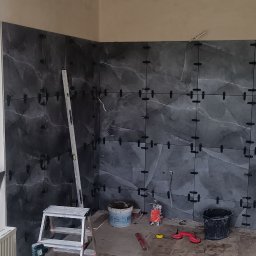 GAMB-POL - Ściana w trakcie układania płytek imitujących marmur, z użyciem krzyżyków dystansowych. Widoczne narzędzia i drabina. Okno z widokiem na zieleń w tle.