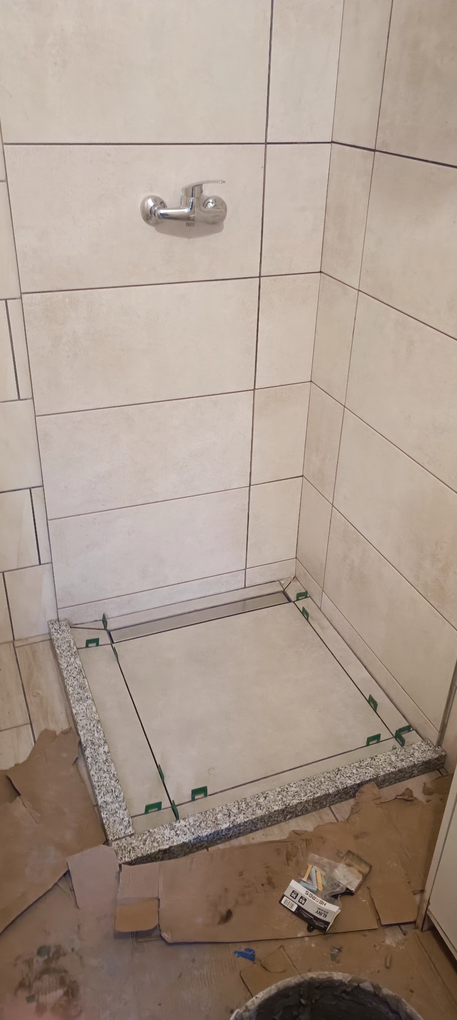 Montaż brodzika w kabinie prysznicowej, widoczne płytki na ścianach, listwa brodzikowa z szarego kamienia, poziomowanie z użyciem klinów, zabezpieczone podłoże kartonem, widoczny kran na ścianie.
