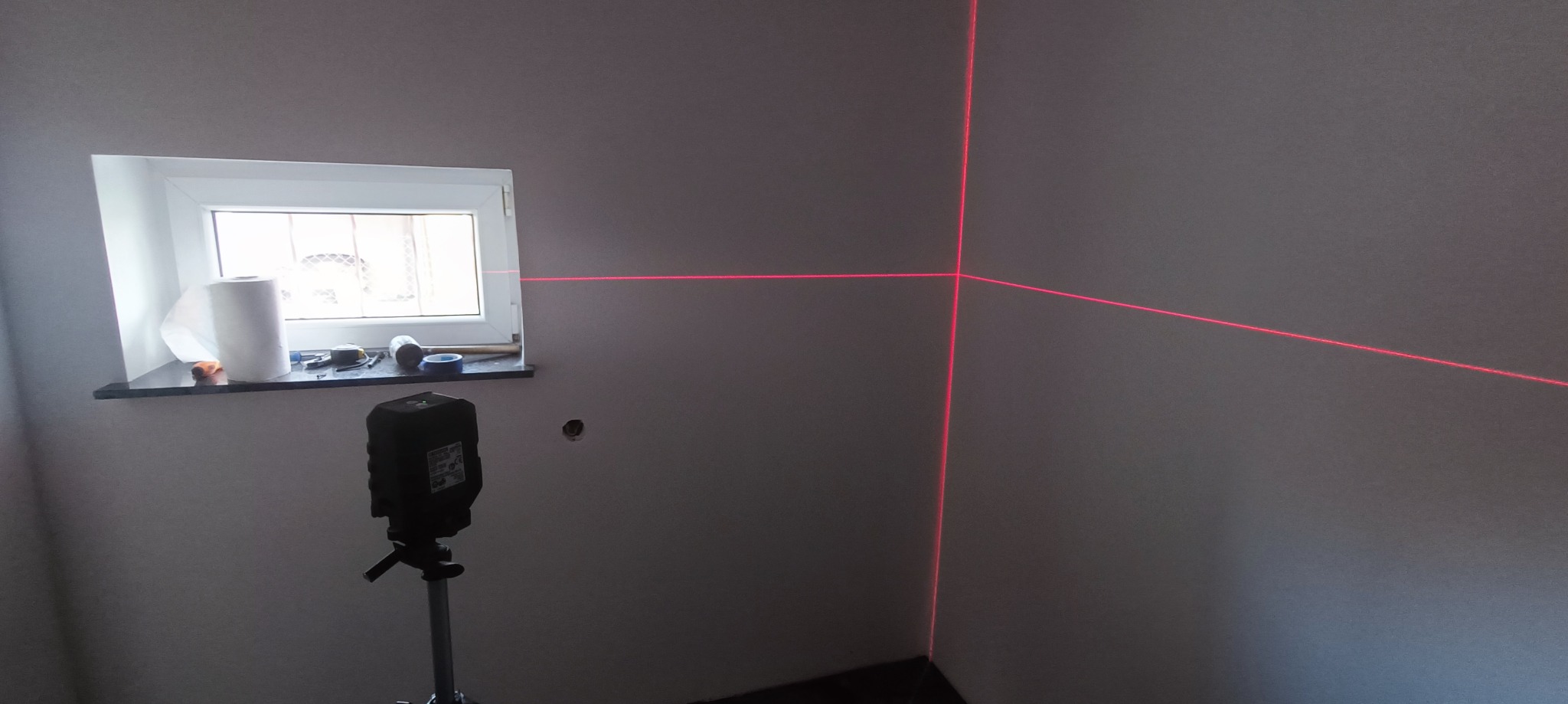 Wnętrze pokoju w trakcie prac wykończeniowych; widoczny laser krzyżowy wyznaczający poziom na ścianach. Na parapecie rolka papieru i narzędzia.