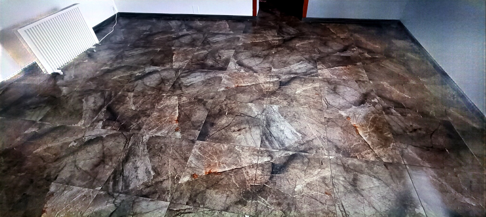 Wykończona ściana z płytek imitujących marmur w odcieniach szarości i brązu, z widocznym grzejnikiem i fragmentem sufitu. Płytki ułożone w regularny wzór.