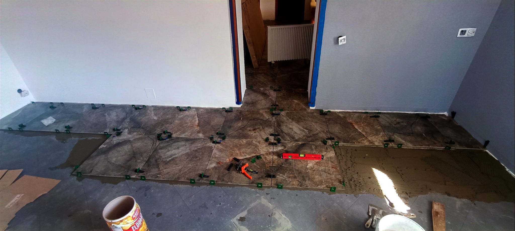 Układanie płytek podłogowych w szaro-brązowej tonacji, z użyciem poziomicy i klipsów poziomujących. Widoczne narzędzia i wiadro. Ściany białe i szare.