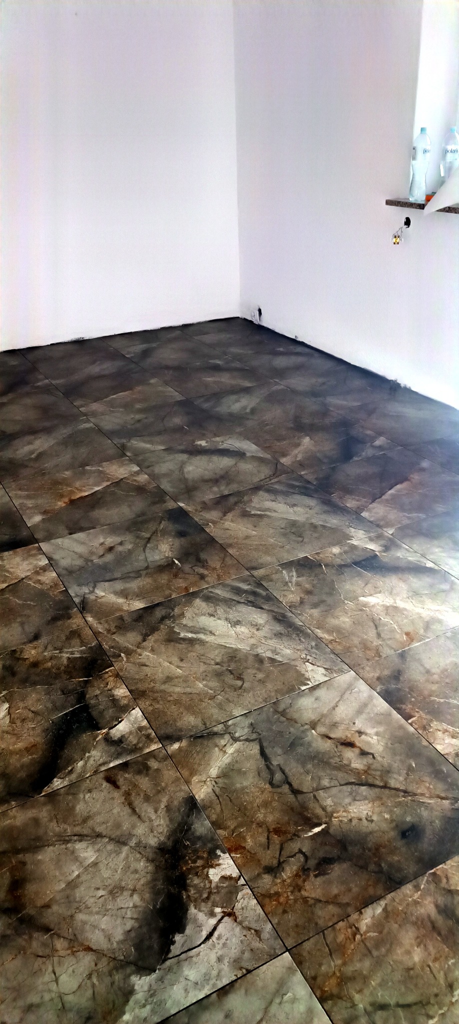 Ukończona podłoga w rogu pokoju wyłożona brązowo-szarymi płytkami imitującymi marmur. Białe ściany i fragment półki z butelkami w tle.
