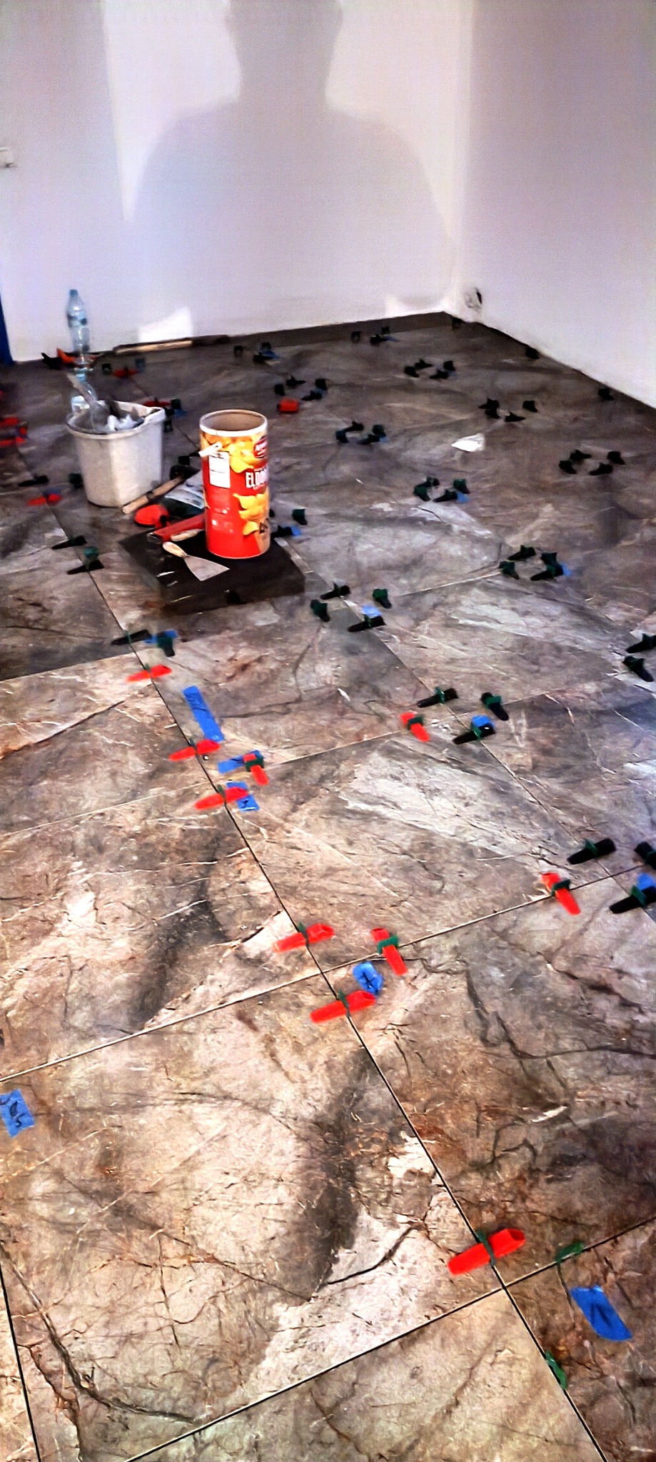 Układanie płytek podłogowych z użyciem systemu poziomowania w Świebodzicach. Widoczne narzędzia, płytki imitujące marmur i cień osoby na ścianie.