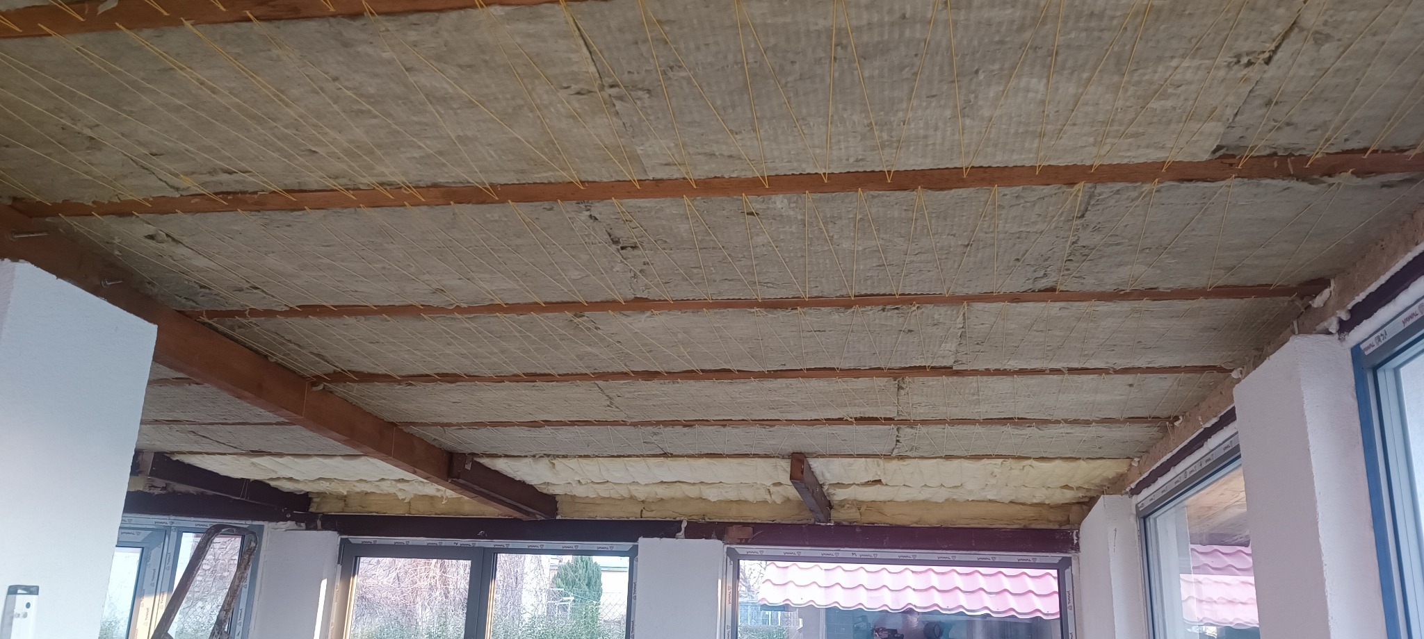 Ocieplenie poddasza wełną mineralną, widoczne drewniane belki stropowe i żółte sznurki mocujące izolację. Prace wykończeniowe w trakcie realizacji.