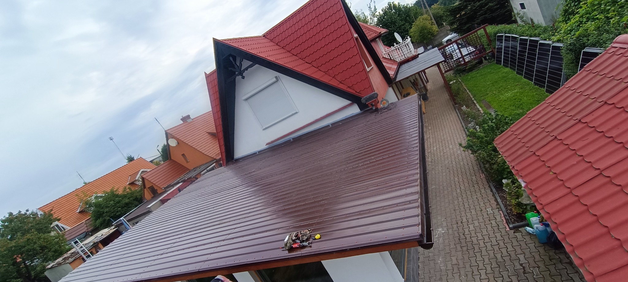 Brązowy dach z blachy trapezowej, widok z góry, narzędzia na dachu, czerwony dach domu w tle, kostka brukowa, zieleń.