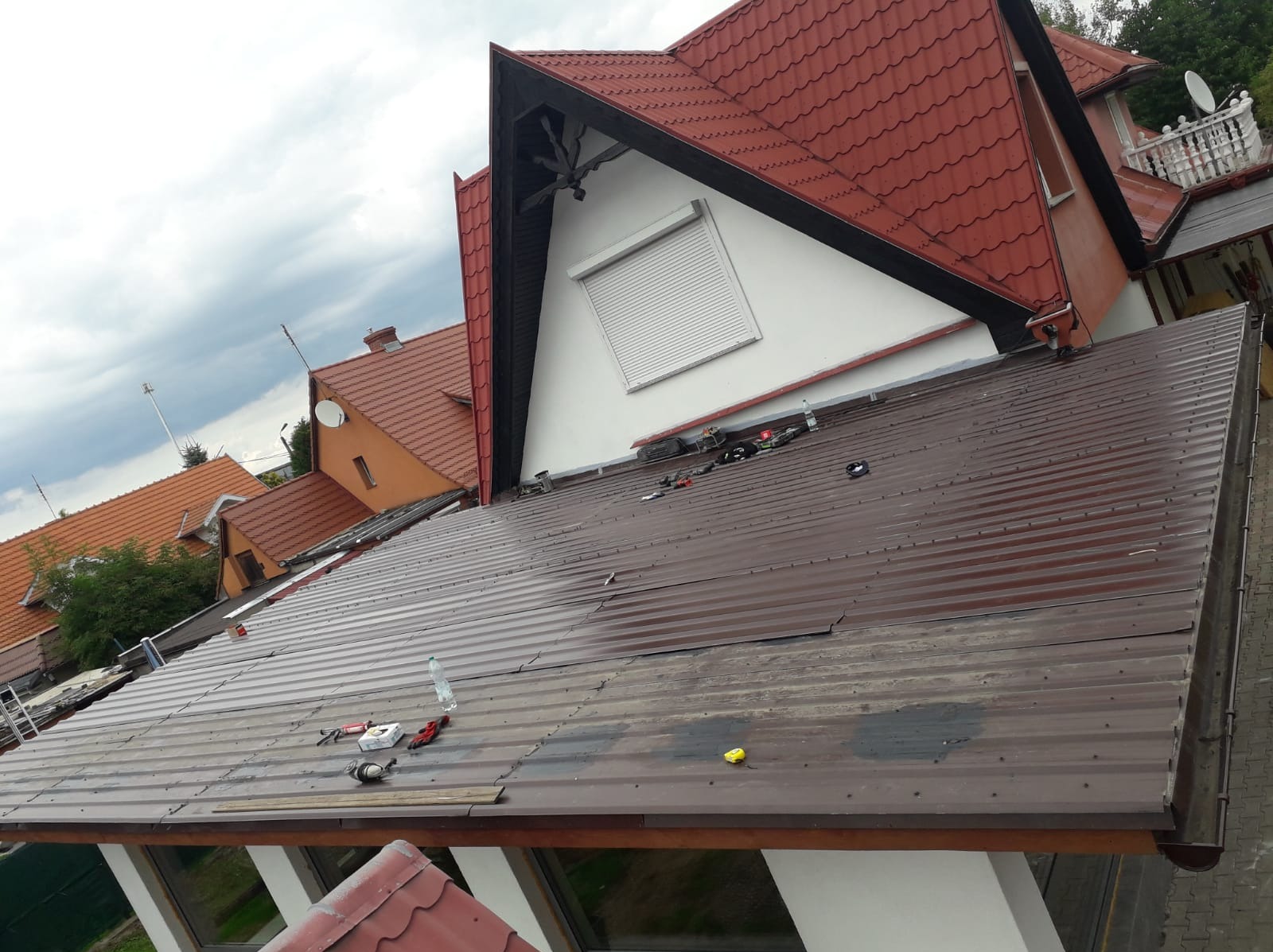 Montaż brązowego dachu z blachy trapezowej na budynku jednorodzinnym. Widoczne narzędzia, częściowo zamontowane panele i fragmenty innych dachów w tle.