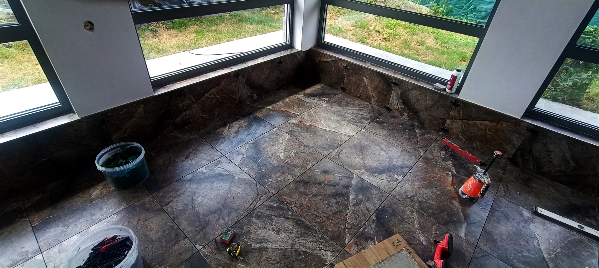 Układanie płytek imitujących marmur w rogu pokoju z oknami. Widoczne narzędzia glazurnicze, poziomica, przyssawka do płytek i pojemniki z akcesoriami.