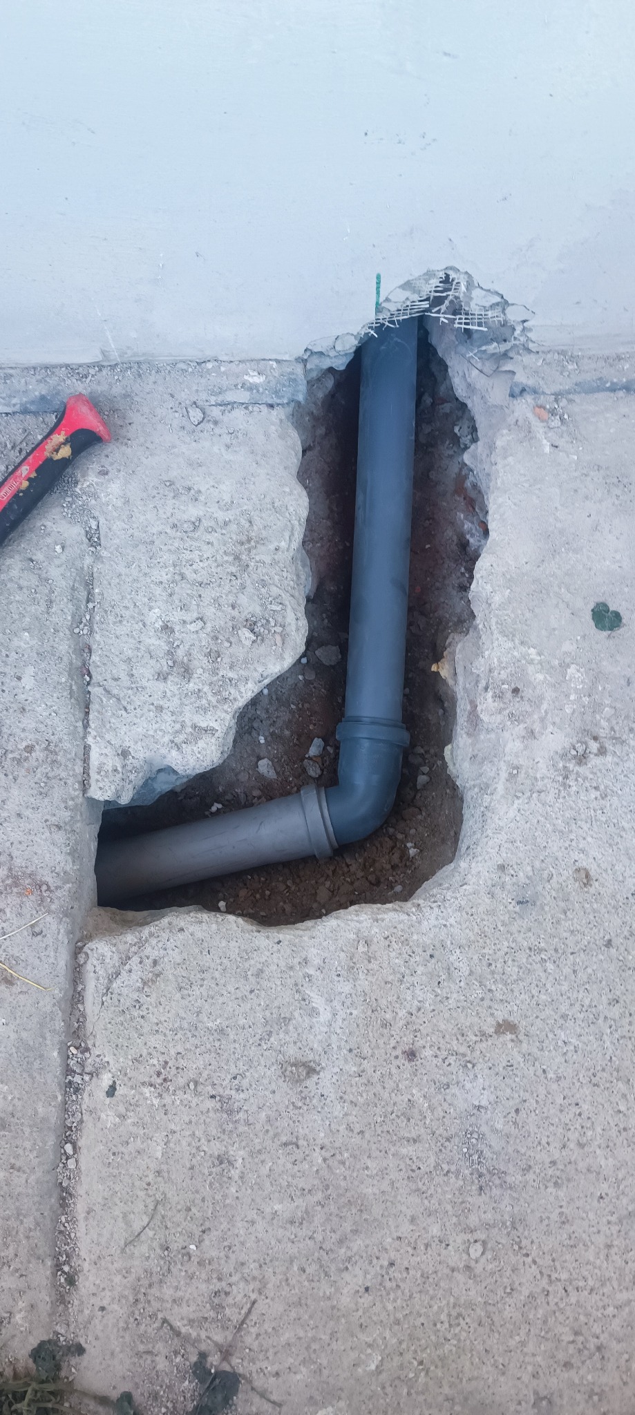 Szare rury kanalizacyjne w betonowej posadzce podczas montażu, widoczny fragment ściany i narzędzie. Instalacja prowadzona pod posadzką.