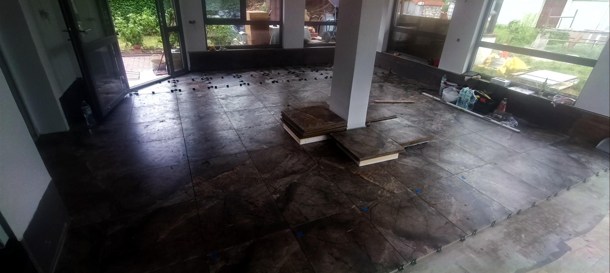 Układanie ciemnych, marmurkowych płytek podłogowych w jasnym pomieszczeniu z dużymi oknami. Widoczne narzędzia i akcesoria glazurnicze, płytki ułożone częściowo.