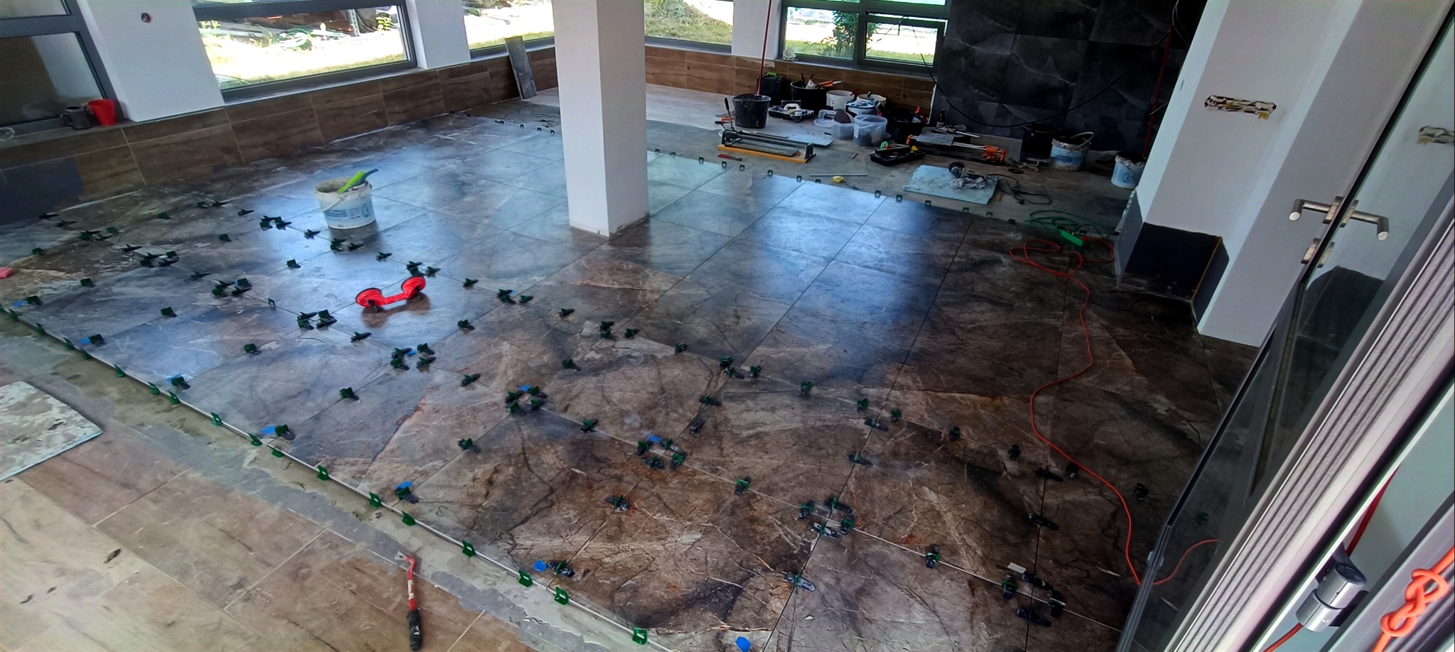 Układanie płytek podłogowych w Świebodzicach. Widoczne narzędzia, poziomice i system poziomowania płytek na brązowych płytkach z marmurowym wzorem w przestronnym wnętrzu.