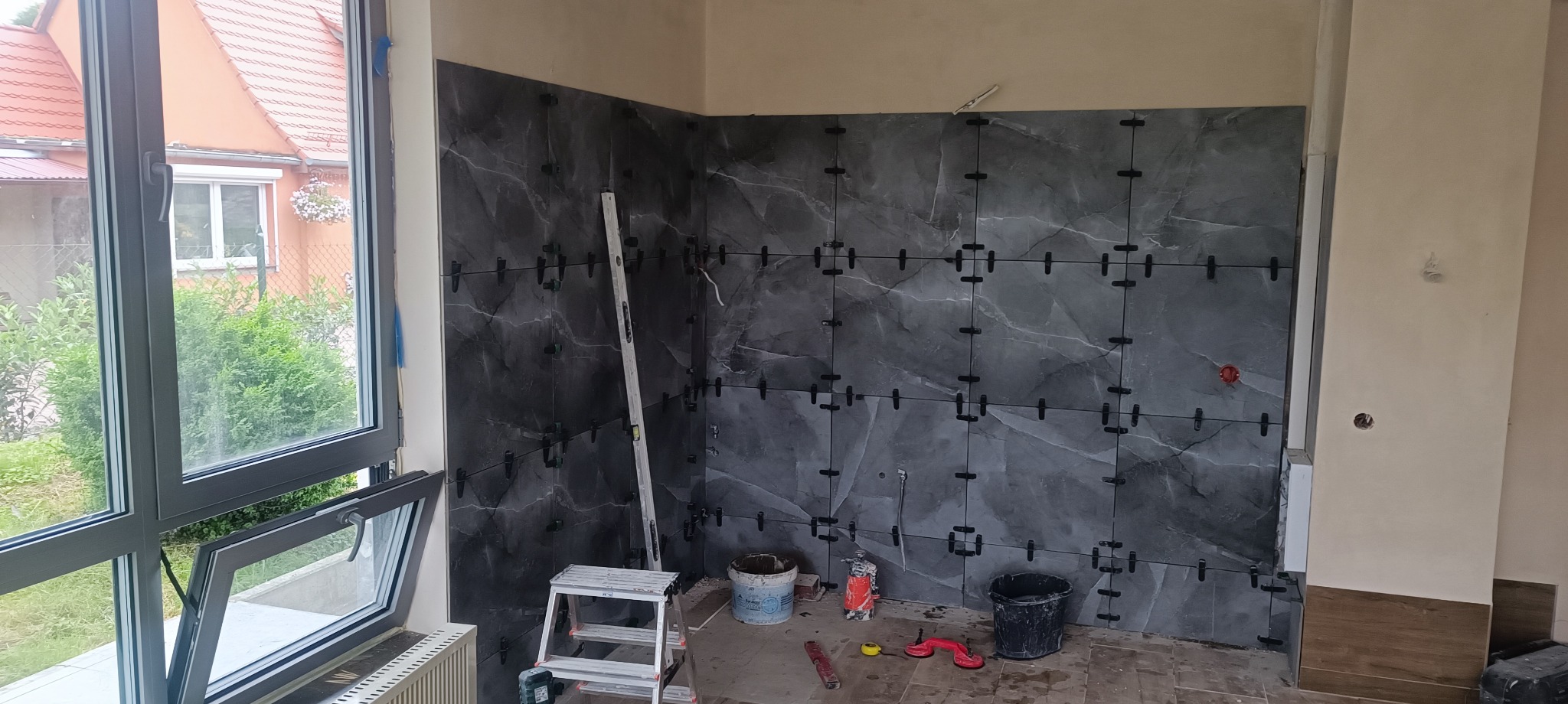 Ściana w trakcie układania płytek imitujących marmur, z użyciem krzyżyków dystansowych. Widoczne narzędzia i drabina. Okno z widokiem na zieleń w tle.