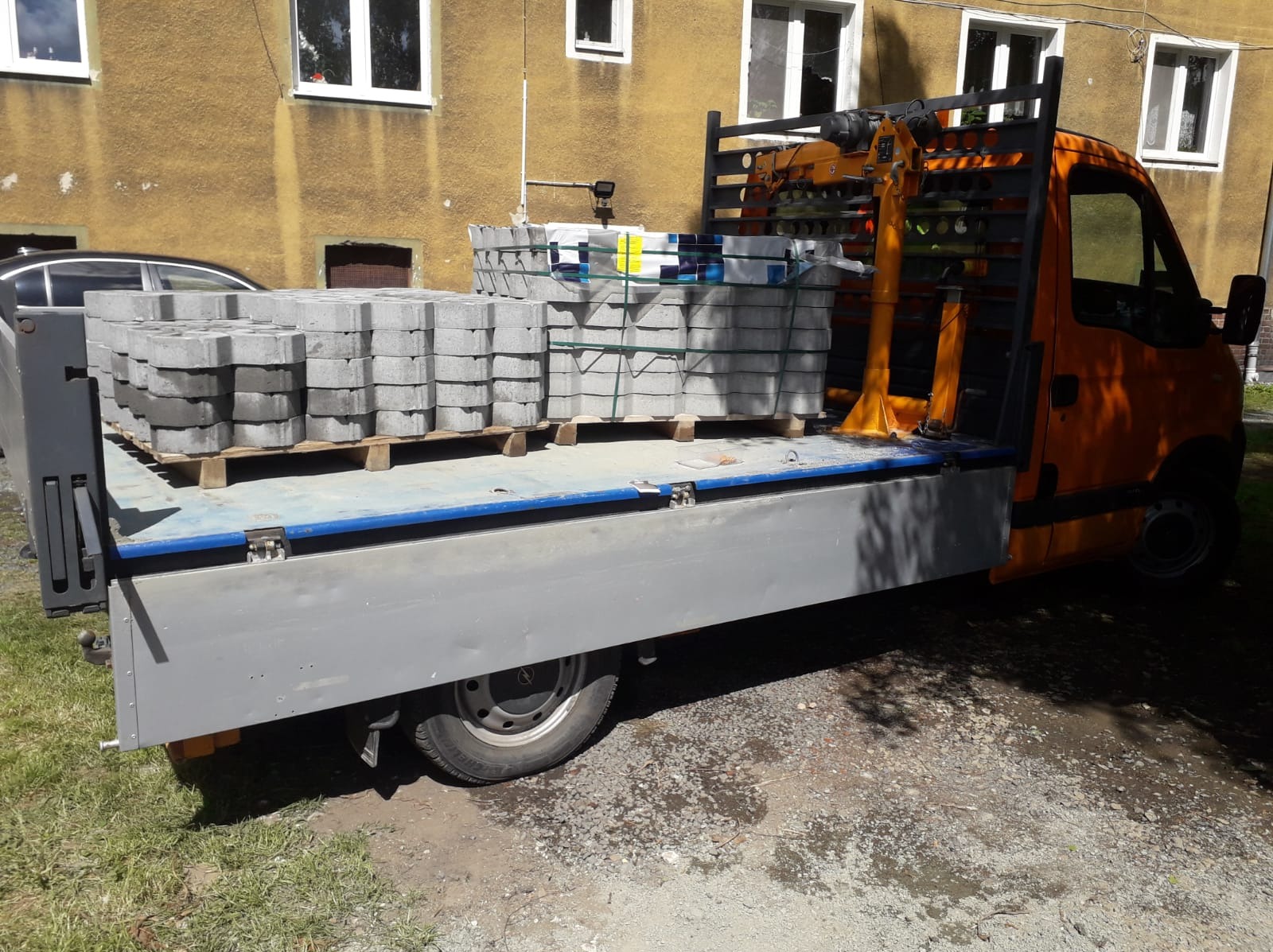 Pomarańczowy samochód dostawczy z paletami szarej kostki brukowej na pace, zaparkowany na trawie przed budynkiem. Widoczny fragment czarnego auta.