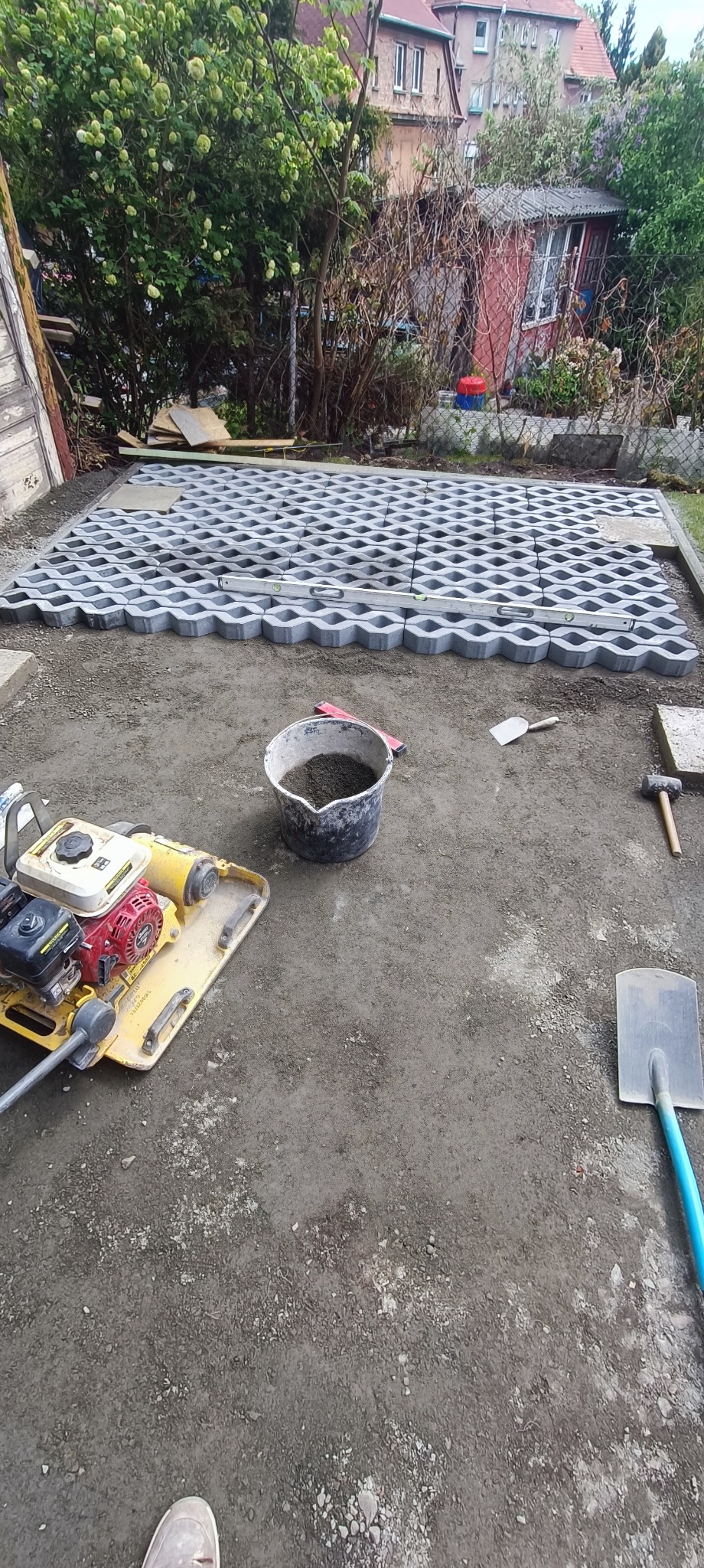 Układanie ażurowych płyt chodnikowych na przygotowanym podłożu. Widoczny ubijak, wiadro z piaskiem i narzędzia. Częściowo ułożona nawierzchnia.