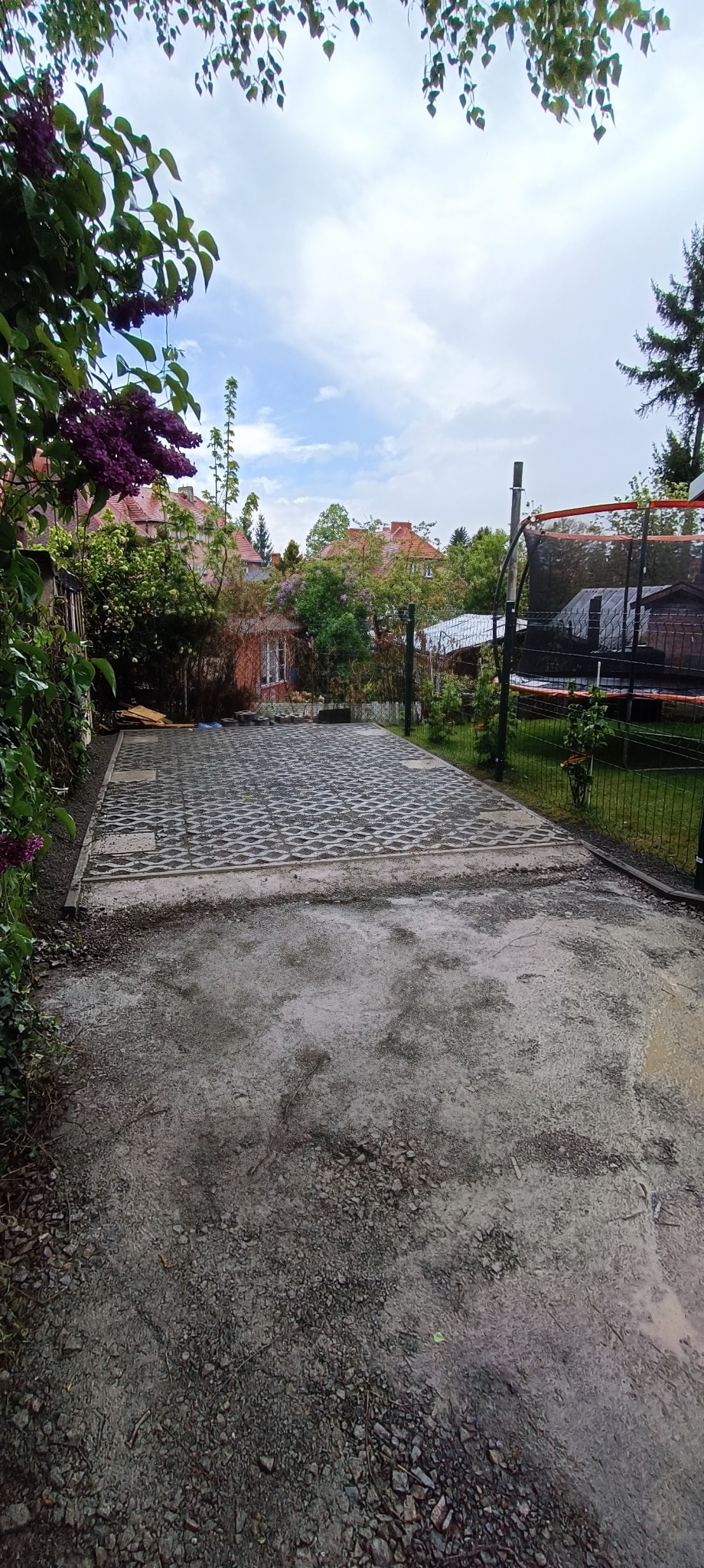 Ułożona szara kostka ażurowa na podwórku, widok z dołu, obok trawnik z trampoliną, w tle budynki mieszkalne i kwitnący bez.