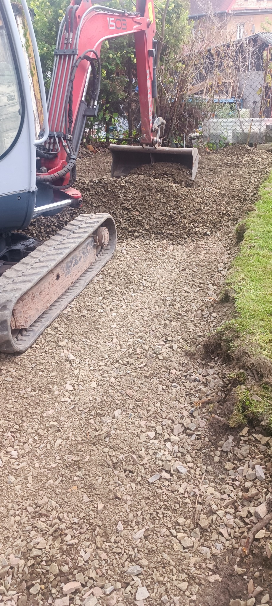 Czerwony mini-koparka wyrównuje teren pod trawnik, widoczne świeżo usunięte krawędzie trawy i przygotowane podłoże z kamieni. Prace ziemne w ogrodzie.