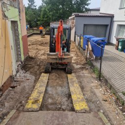 MP TECH - Mała koparka na żółtych najazdach na tle placu budowy i częściowo rozebranego budynku.