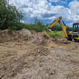 MP TECH - Żółta koparko-ładowarka New Holland podczas prac ziemnych, operator w kabinie, wykopany dół w ziemi, w tle drzewa i błękitne niebo z chmurami.