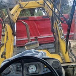 MP TECH - Widok z kabiny brudnej koparko-ładowarki na czerwoną wywrotkę, załadowaną ziemią i gałęziami, widoczne elementy sterujące i kierownica.