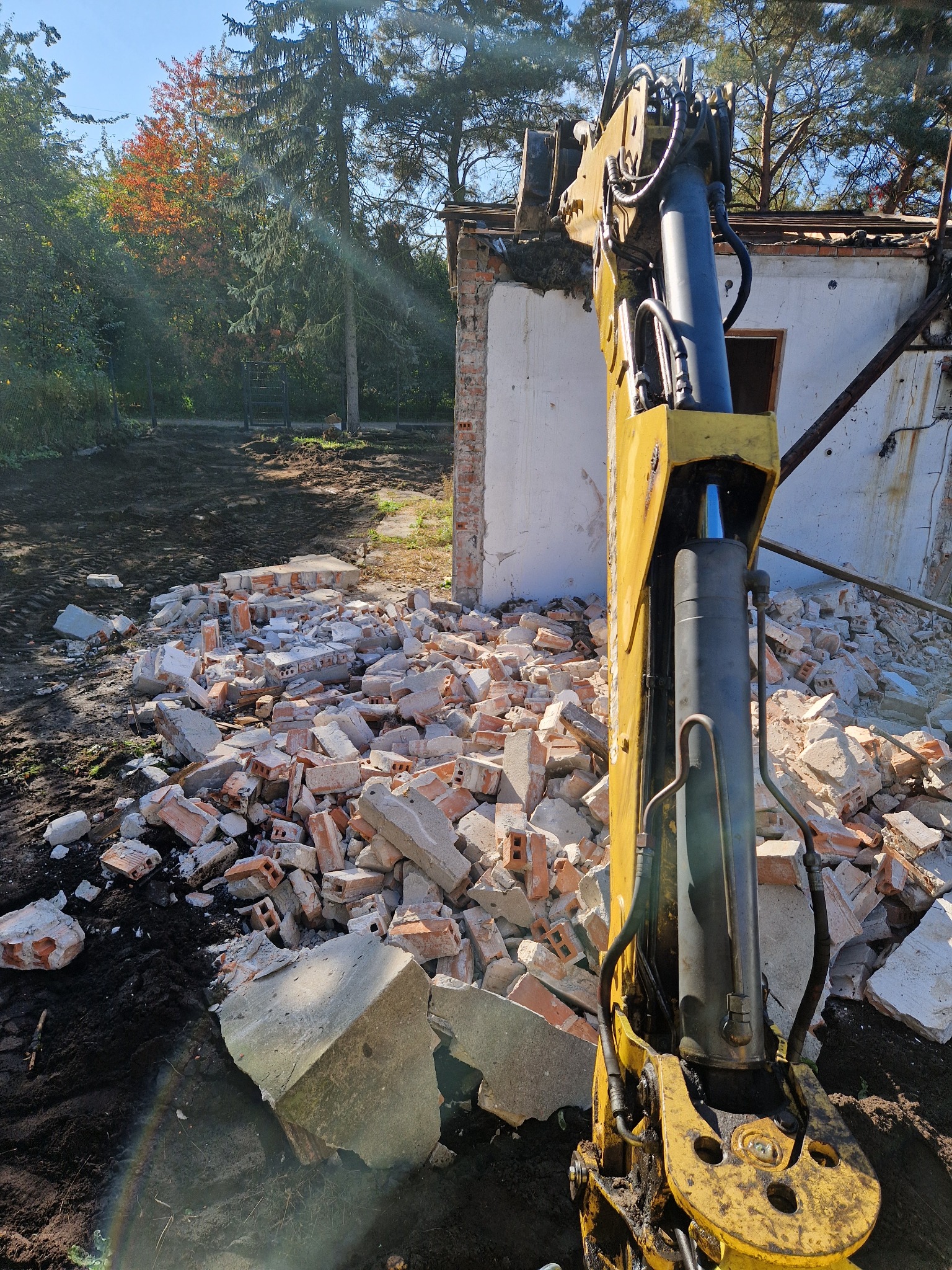 Żółty hydrauliczny młot wyburzeniowy na tle gruzowiska i resztek białego budynku, częściowo zburzonego, z zielenią w tle.