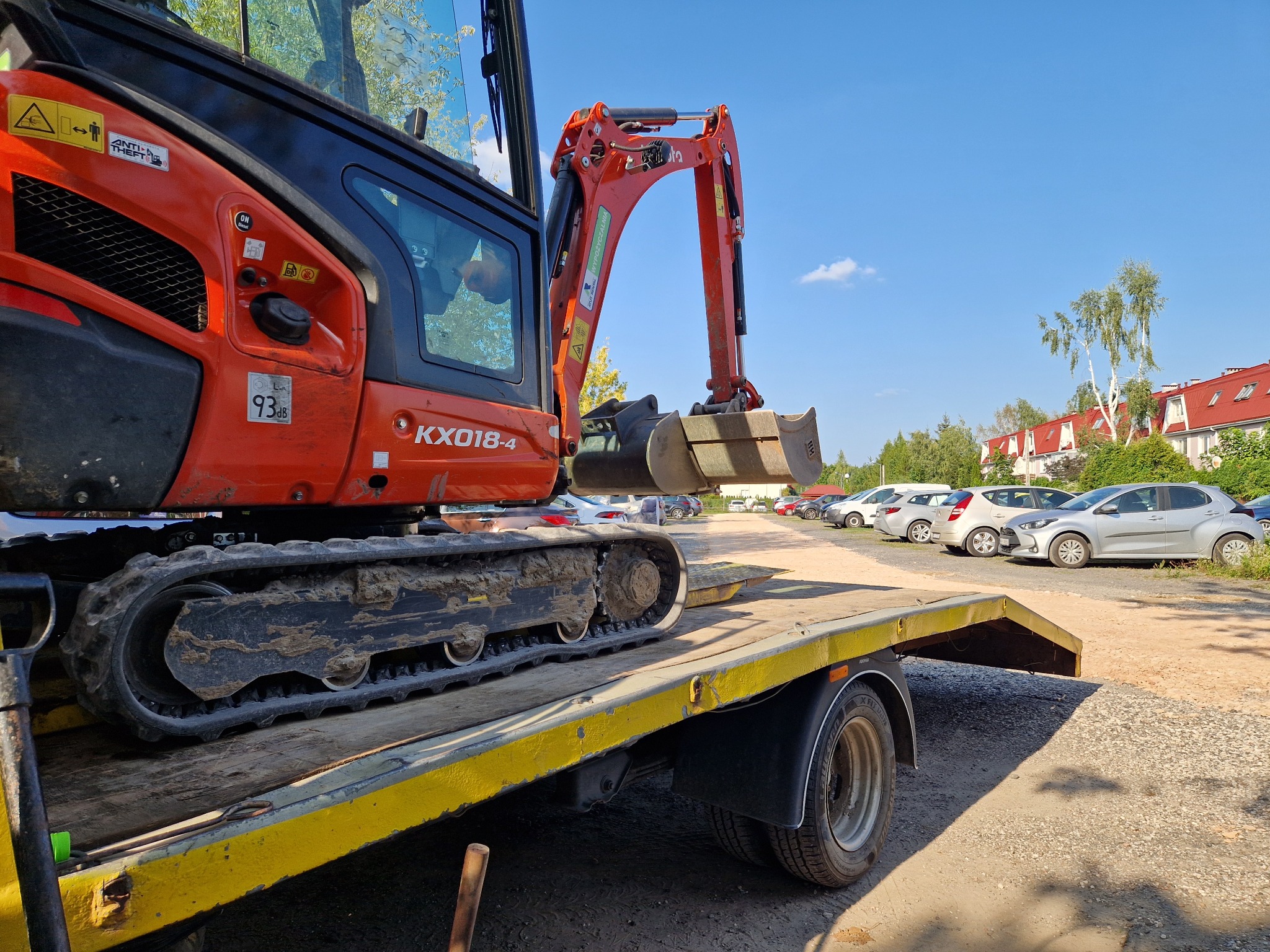 Pomarańczowa minikoparka Kubota KX018-4, z ubłoconymi gąsienicami, załadowana na żółtą lawetę. W tle parking z samochodami i budynki mieszkalne.