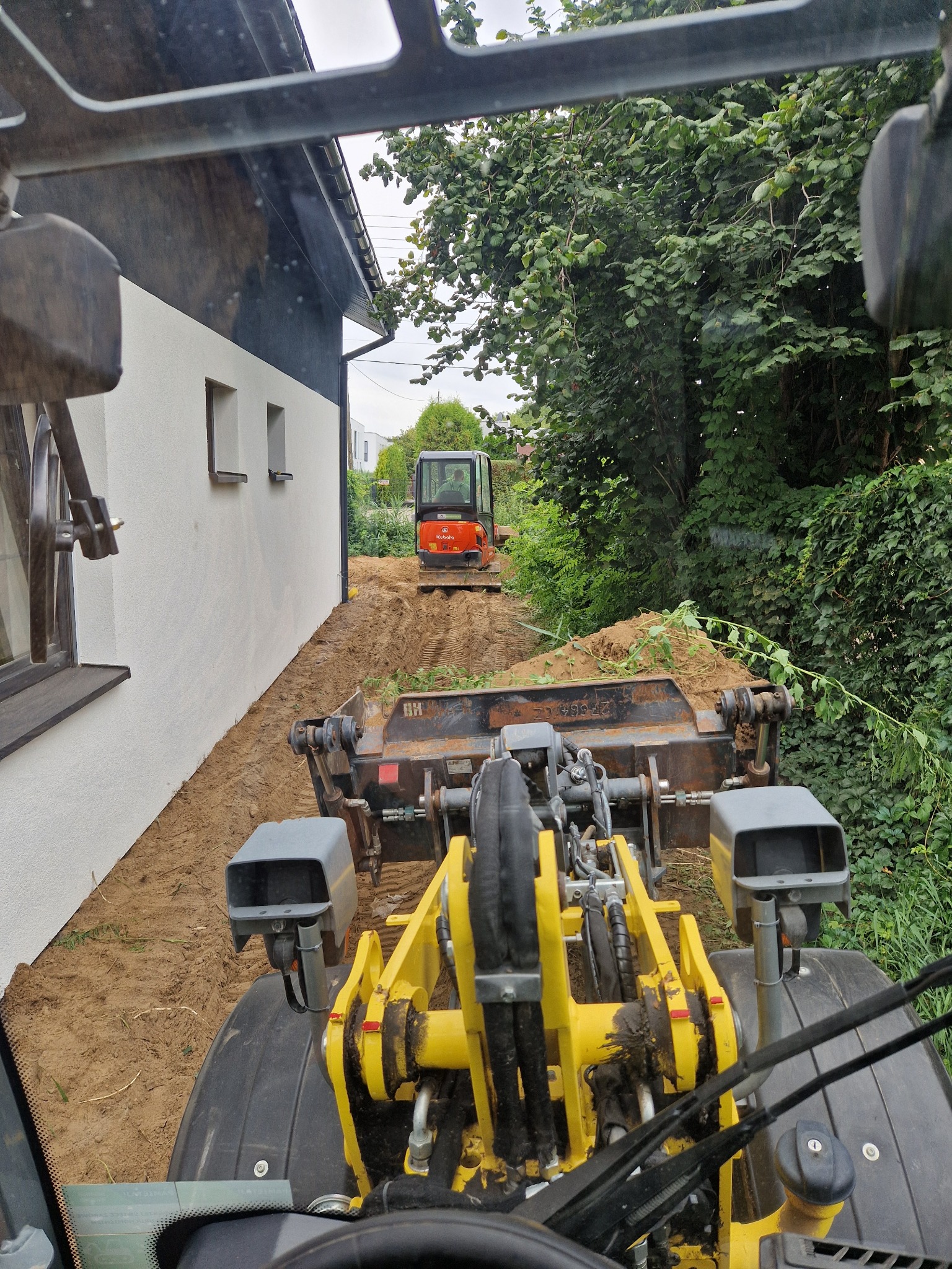 Widok z kabiny żółtej ładowarki na pomarańczową mini-koparkę pracującą w wykopie wzdłuż białego budynku z małymi oknami.