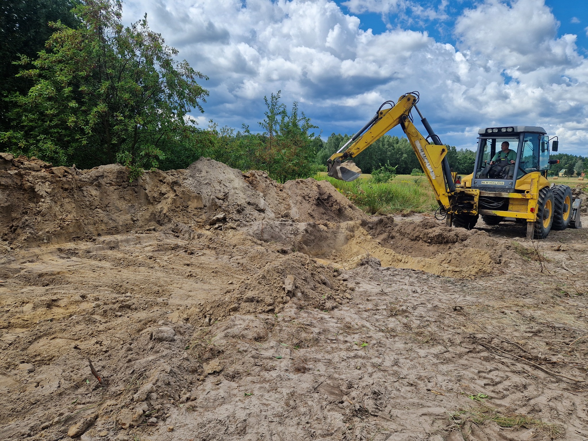 Żółta koparko-ładowarka New Holland podczas prac ziemnych, operator w kabinie, wykopany dół w ziemi, w tle drzewa i błękitne niebo z chmurami.