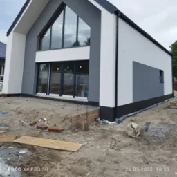 Nowoczesny dom z elewacją w odcieniach szarości i bieli, duże okna, teren wokół w trakcie prac wykończeniowych. Widoczne materiały budowlane.