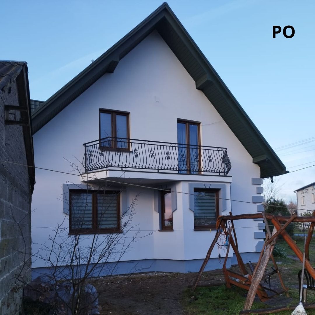 Dwupiętrowy dom z białymi ścianami, ciemnymi oknami i balkonem z kutą balustradą, widoczny z boku w pochmurny dzień, z huśtawką na pierwszym planie.