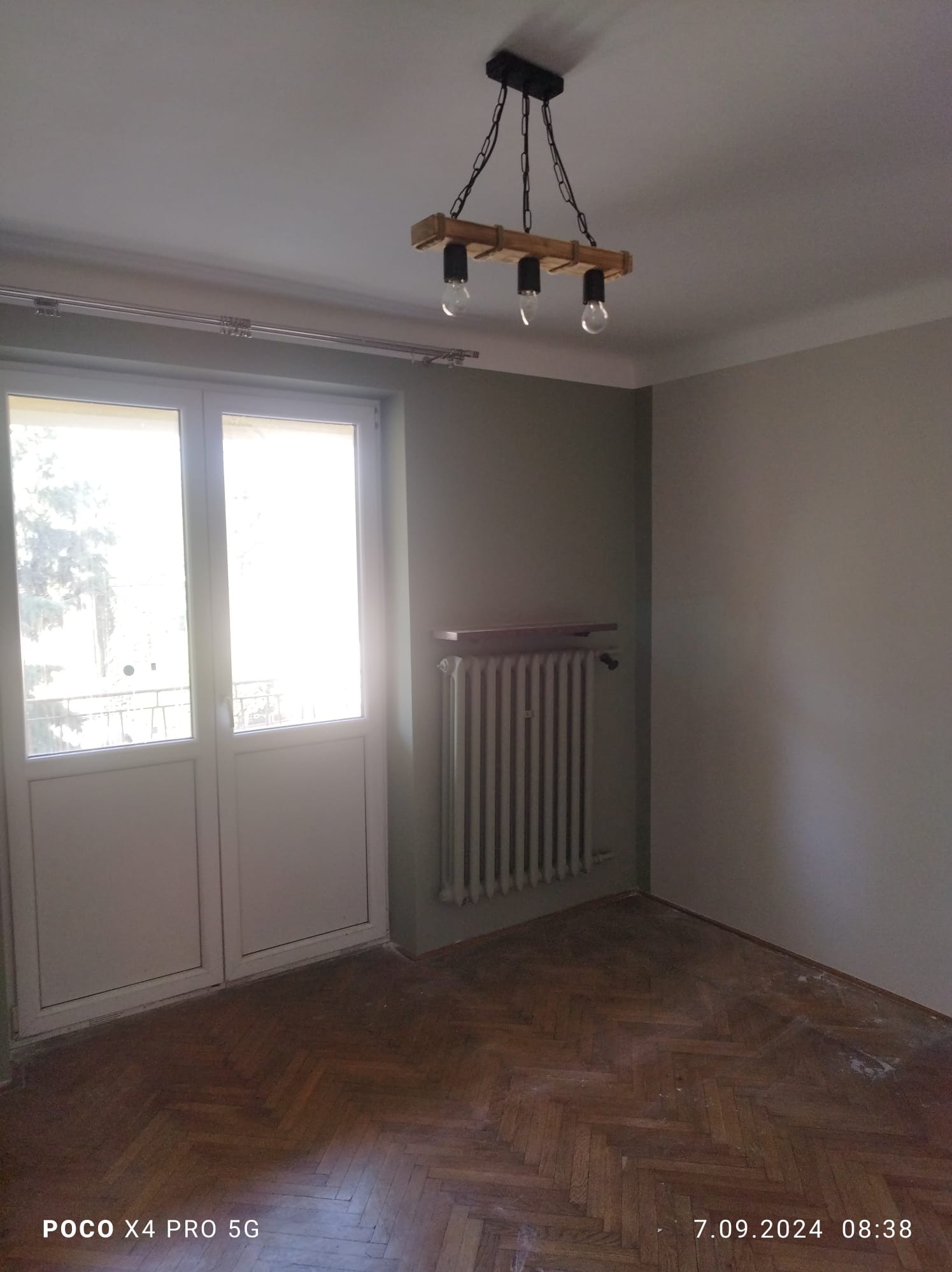 Pomalowane na szaro wnętrze pokoju z drewnianą podłogą w jodełkę, grzejnikiem pod parapetem i nowoczesną lampą z drewnianymi elementami. Widoczne ślady po remoncie na podłodze.