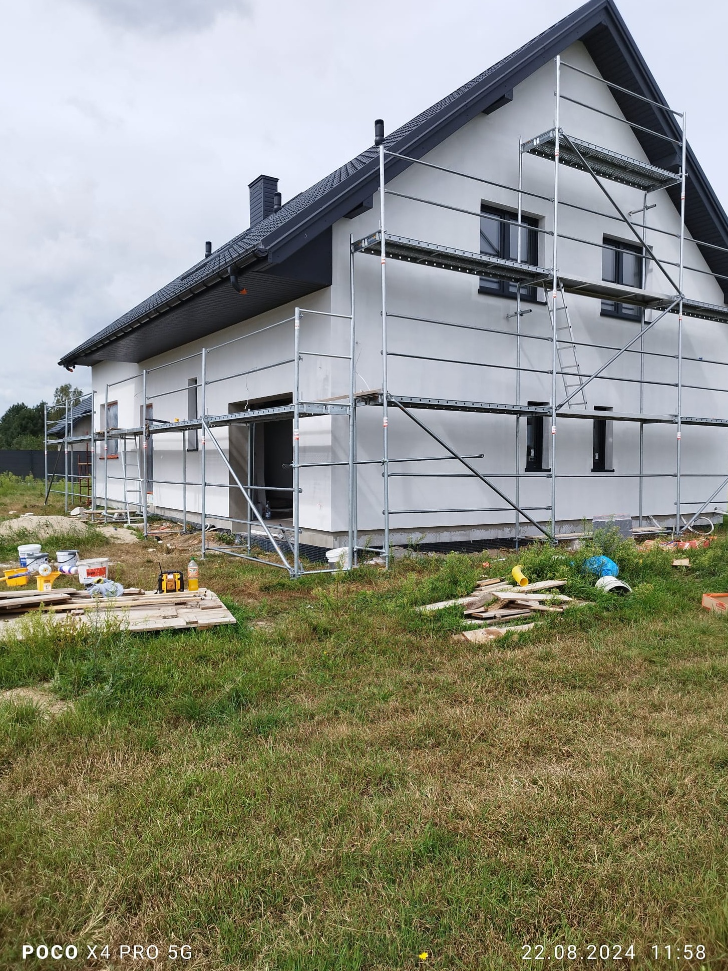 Dom jednorodzinny z jasną elewacją w trakcie prac, otoczony rusztowaniem, z widocznymi narzędziami i materiałami budowlanymi na trawie.