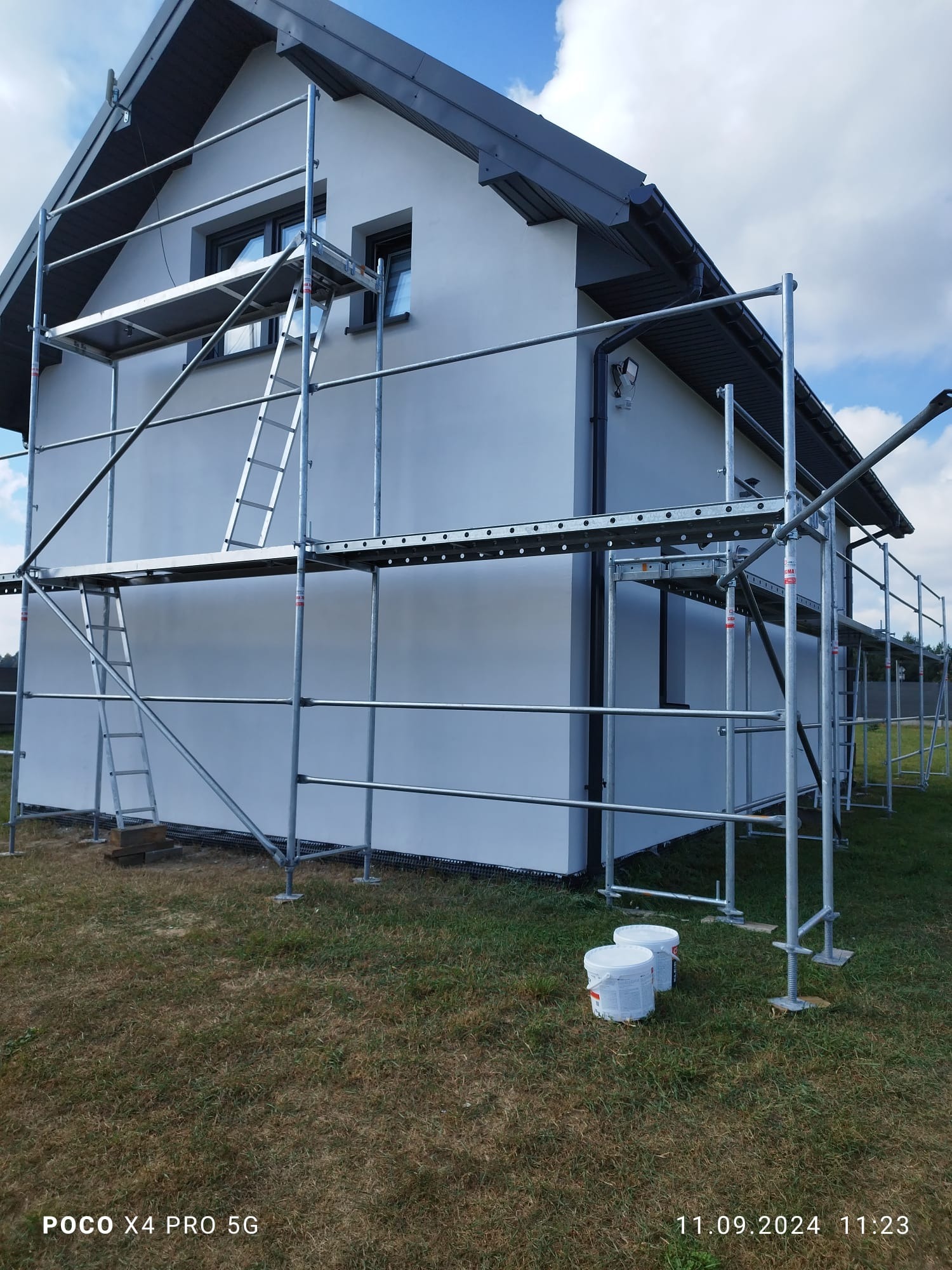 Dom jednorodzinny z jasną elewacją w trakcie ocieplania, widoczne rusztowania i dwa białe wiadra z farbą u podstawy ściany.