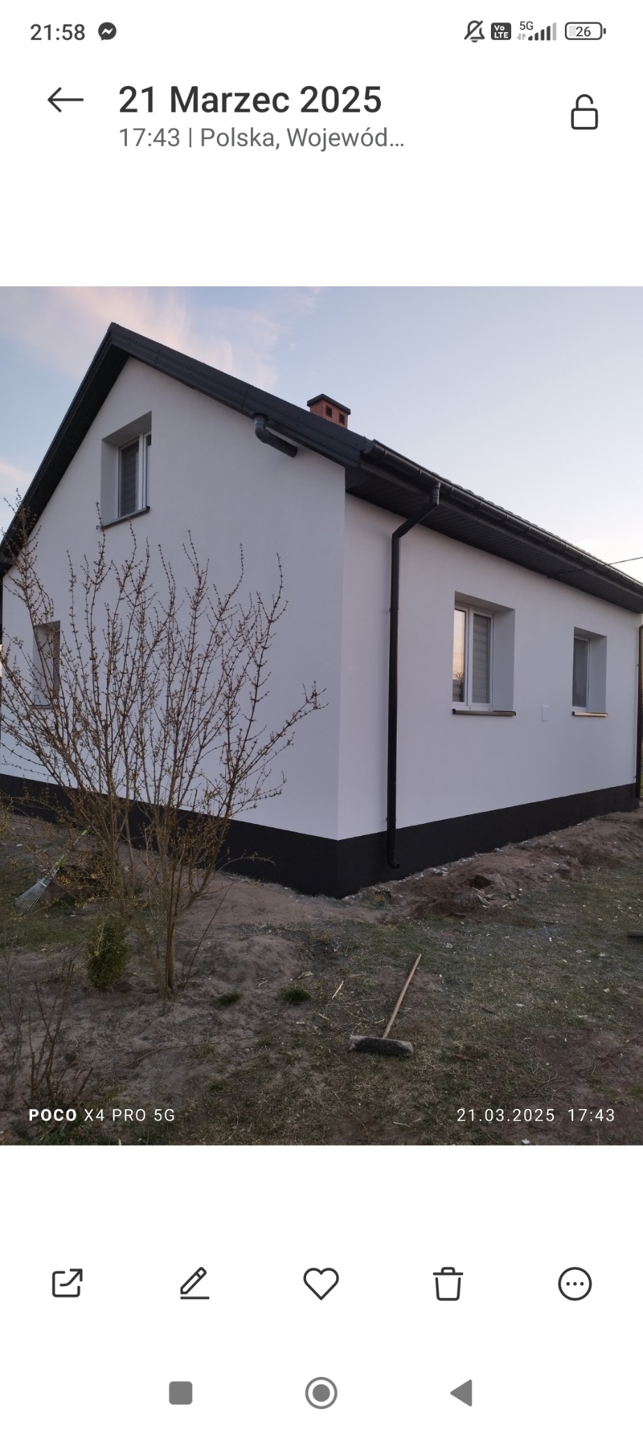 Biały dom jednorodzinny z czarnym dachem i fundamentami, widoczny krzew bez liści i leżąca zmiotka.