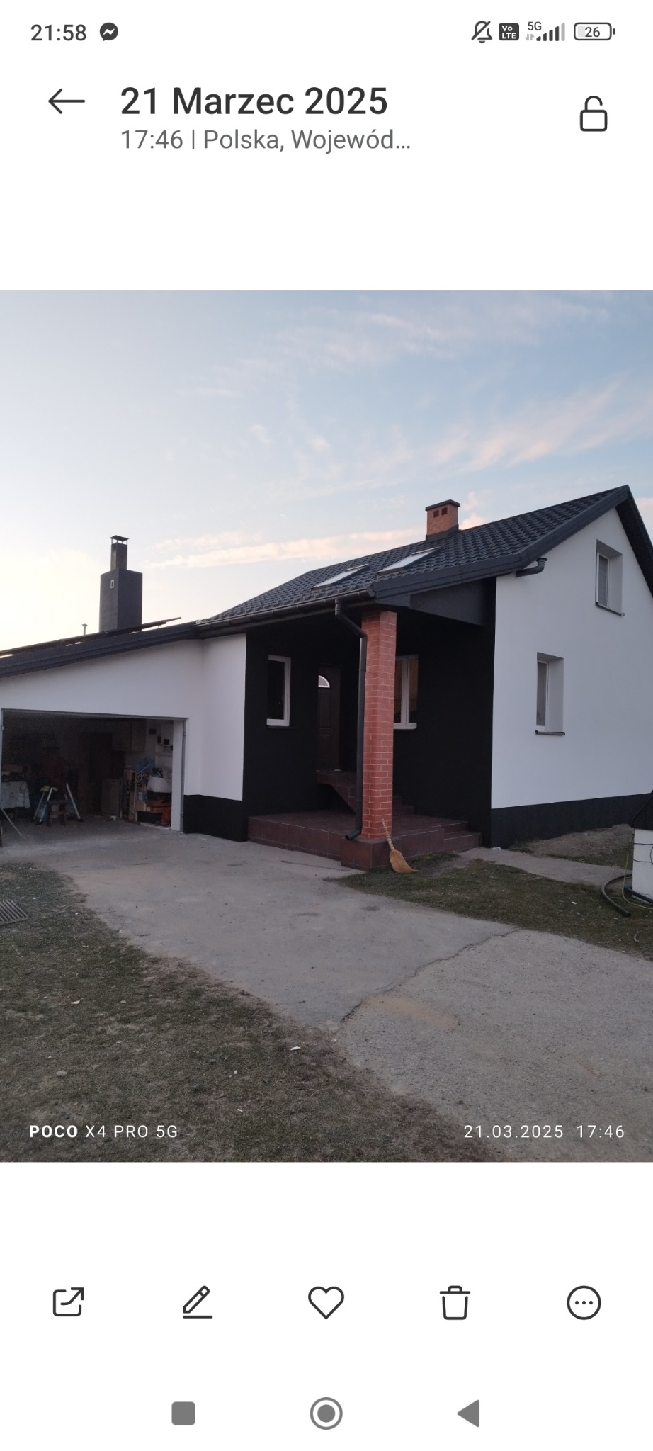 Dom jednorodzinny z czarną elewacją i białymi elementami, widoczny z zewnątrz, z otwartym garażem i częściowo wybrukowanym podjazdem, w tle błękitne niebo.