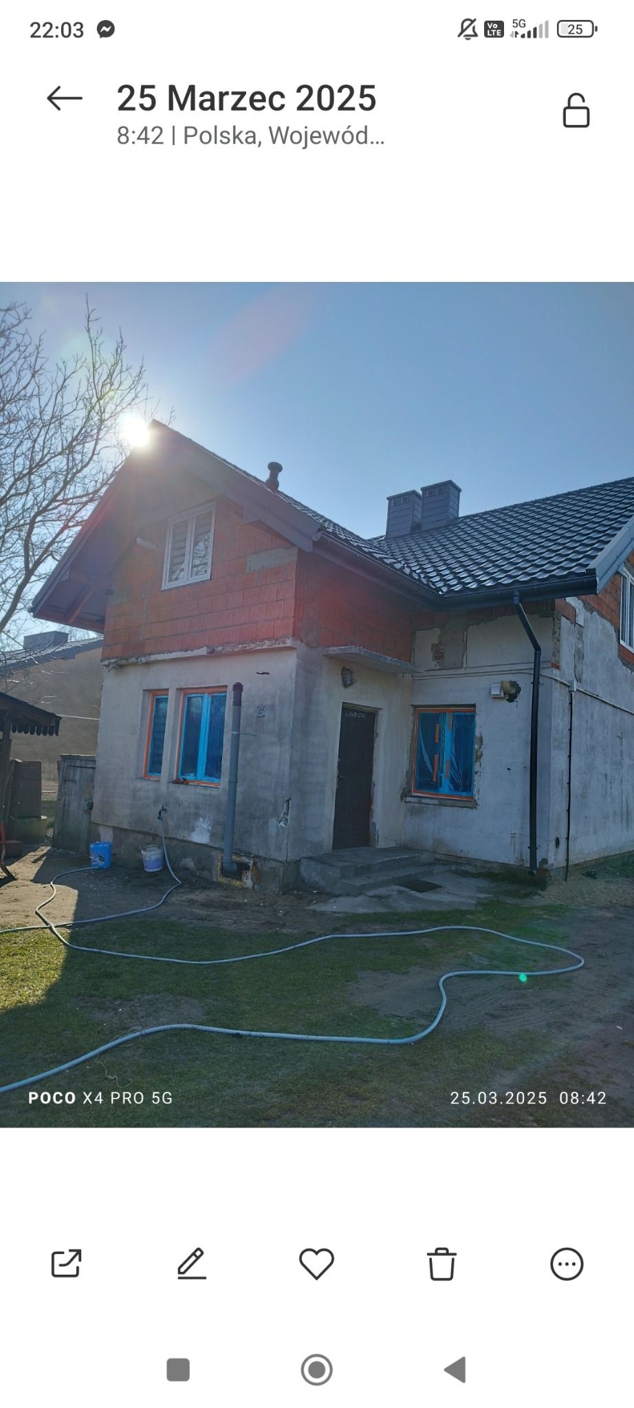 Dom w trakcie prac budowlanych: część elewacji z cegły, okna zabezpieczone folią, widoczne ślady zaprawy, na trawniku leży wąż ogrodowy.