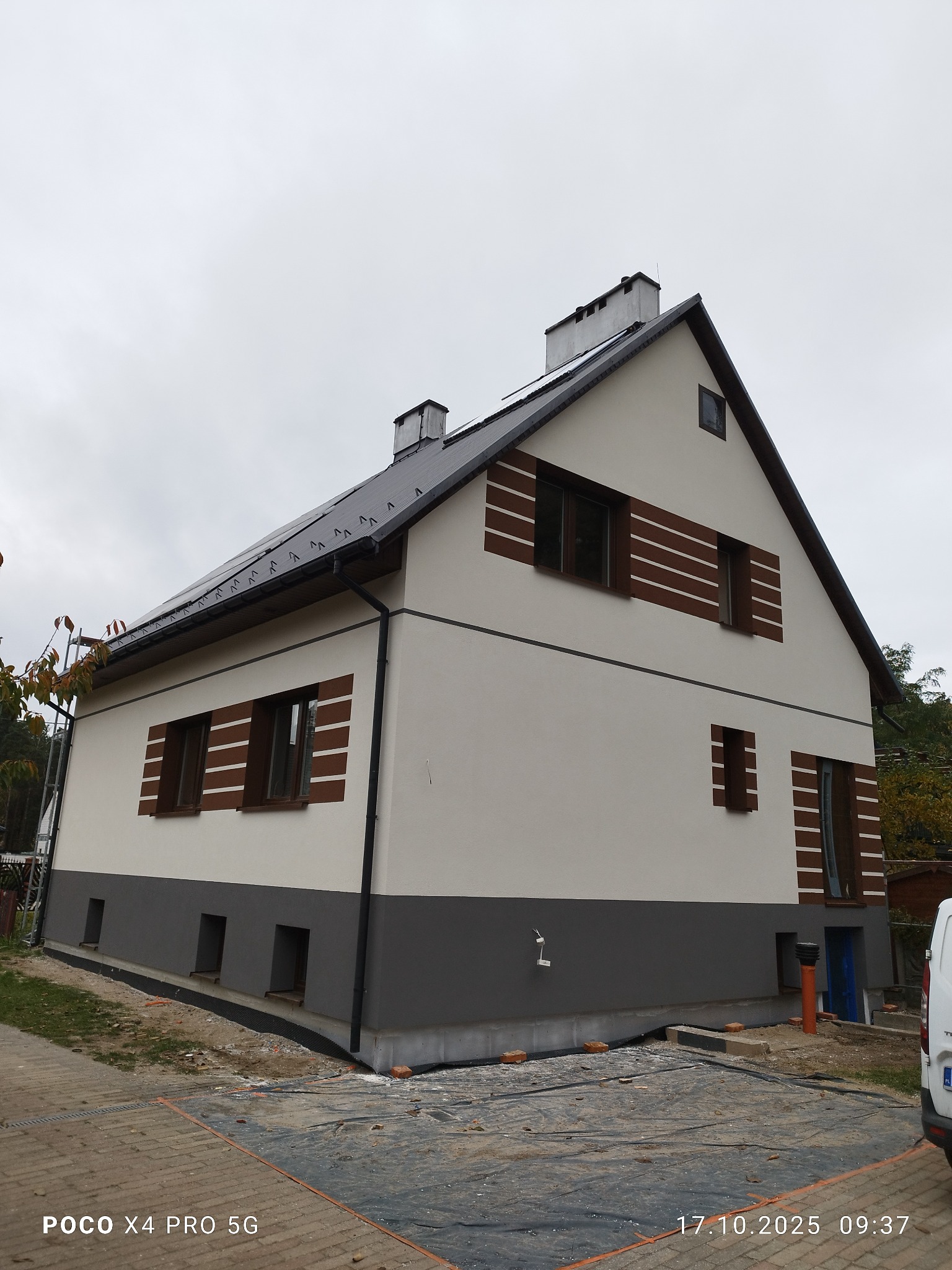 Dwukondygnacyjny dom po renowacji elewacji z dekoracyjnymi, poziomymi pasami w kolorze brązu wokół okien. Dolna część elewacji w odcieniu szarości.