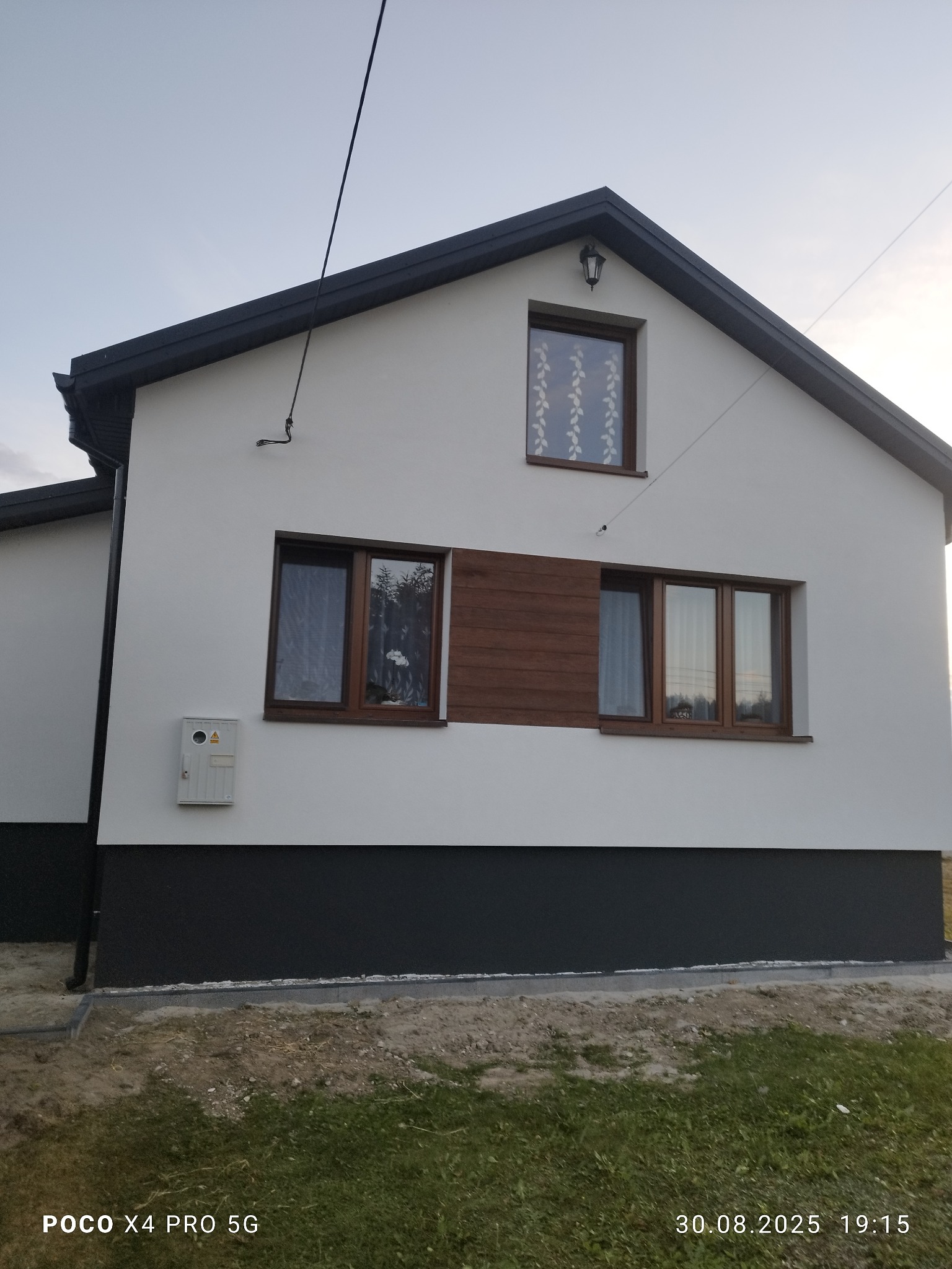 Biała elewacja domu jednorodzinnego z ciemną podmurówką i drewnianą wstawką obok okna. Widoczne okna z firankami i lampa nad oknem na poddaszu.