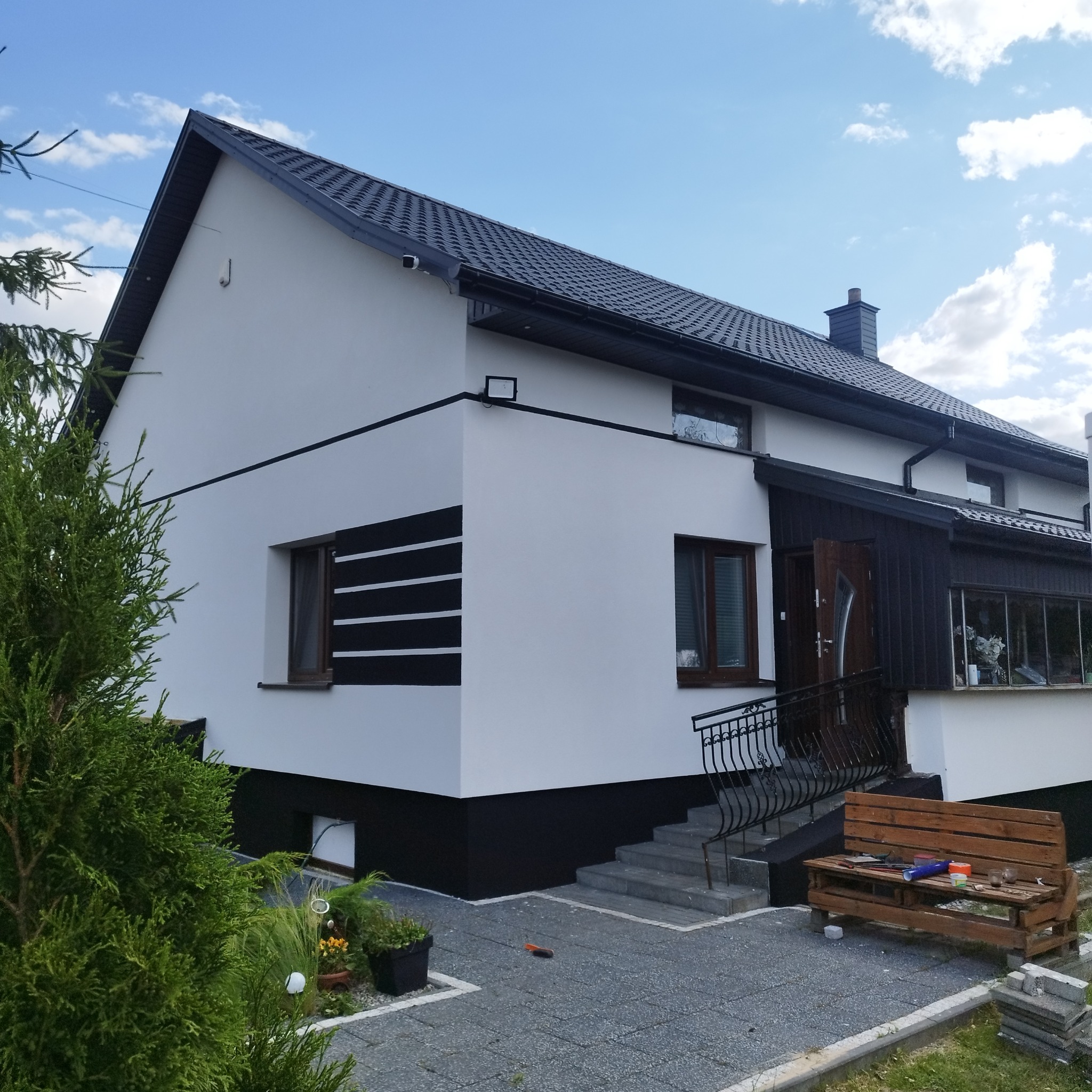 Biała elewacja domu z czarnymi akcentami: pasy dekoracyjne, podmurówka i dach. Widoczne schody wejściowe, fragment ogrodu i narzędzia malarskie na ławce.
