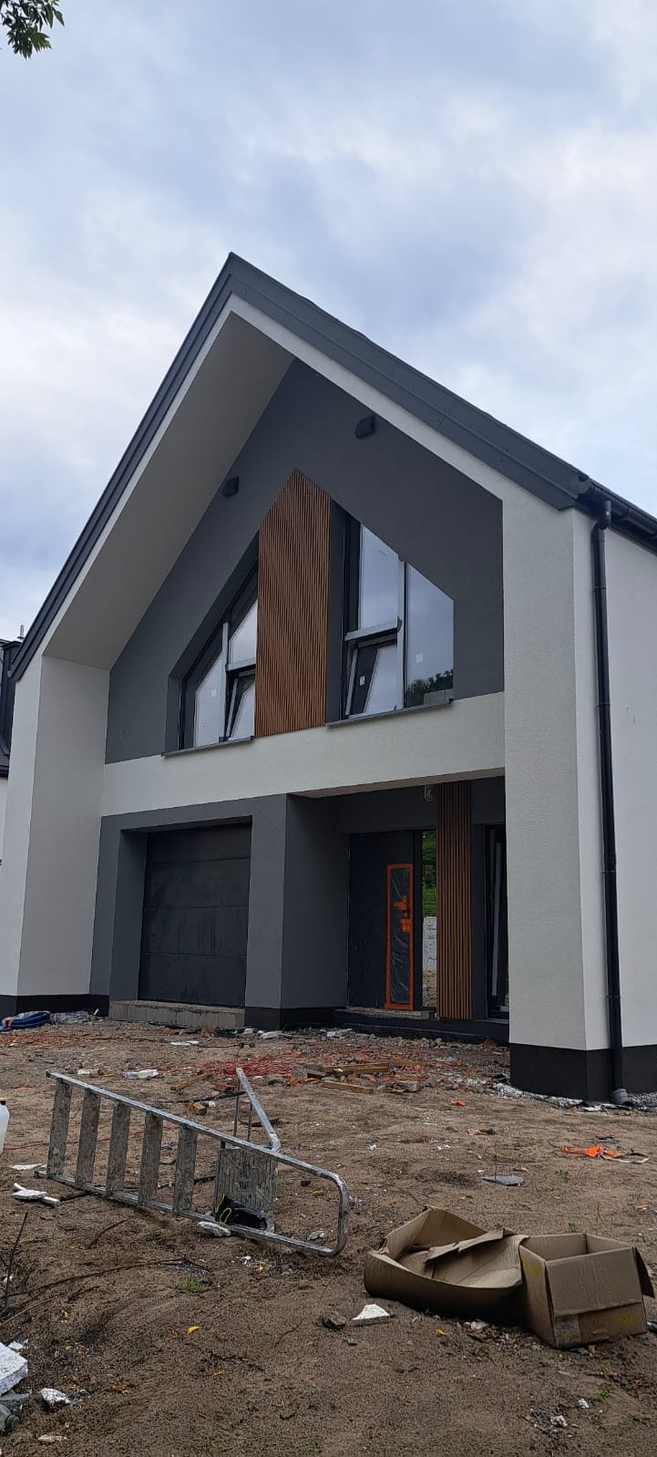 Nowoczesny dom jednorodzinny z szarą elewacją i drewnianymi panelami dekoracyjnymi. Widoczne okna o nietypowym kształcie i brama garażowa. Teren wokół domu w trakcie prac wykończeniowych.