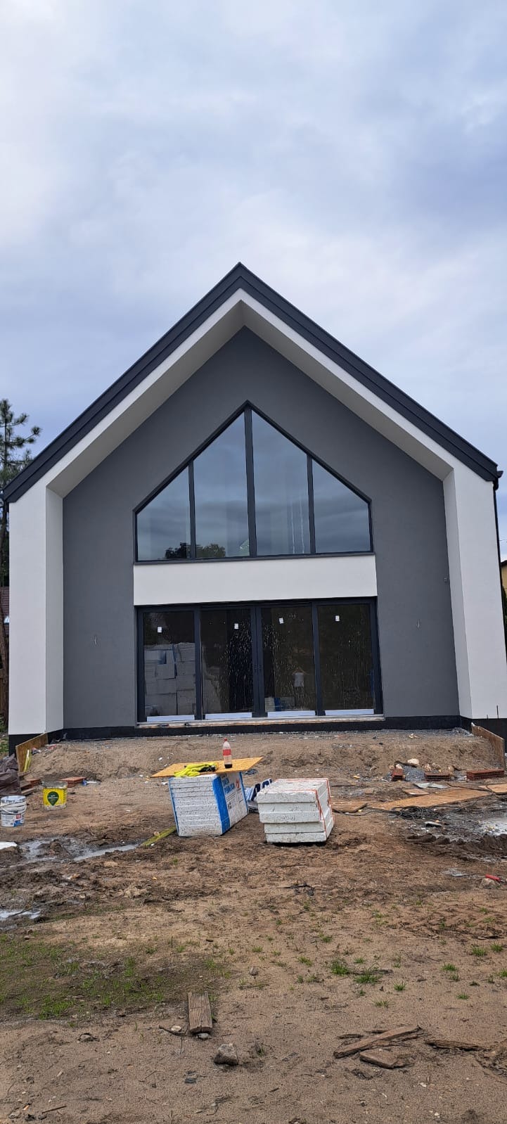 Nowoczesny dom jednorodzinny z dużymi oknami, świeżo otynkowany na szaro i biało, z widocznymi materiałami budowlanymi na posesji. Minimalistyczny design.