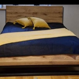 Holleywood - Łóżko z drewnianym wezgłowiem i ramą na metalowych nogach, przykryte granatową narzutą i żółtymi poduszkami, w sypialni.