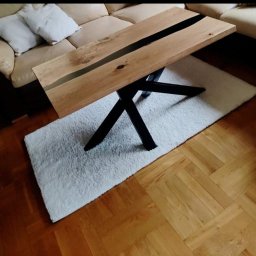 Holleywood - Stolik kawowy z blatem z jasnego drewna z czarną żywicą epoksydową wzdłuż środka, na czarnych, metalowych nogach w kształcie litery X, na białym dywaniku typu shaggy, na parkiecie.