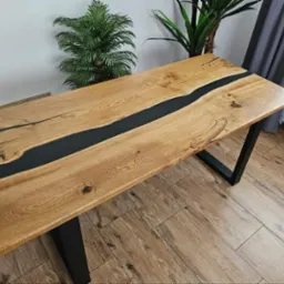 Blat stołu z drewna z żywicą epoksydową w kolorze czarnym, osadzony na metalowych nogach w kształcie trapezu, na tle roślin doniczkowych i podłogi z paneli imitujących drewno.