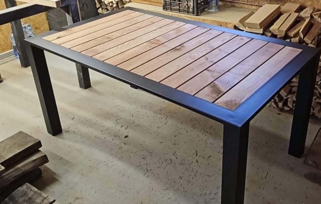 Stół z drewnianym blatem ułożonym z desek i metalową, czarną ramą, widoczny w warsztacie stolarskim z zapasem drewna w tle.